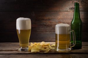 Two mugs of beer with thick foam heads sit adjacent to an almost-empty green bottle and a pile of potato chips against a woodgrain background.