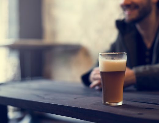 Slightly out of focus young man with closely trimmed beard sits at a wooden picnic table indoors with a craft beer.
