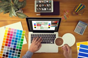 POV shot shows color swatches, a laptop with a design program selected, a tablet with a corresponding program, and various office products on the desk like colored paperclips, a plant, a calculator, and a colorful arrangement of pencils in a glass.. A pair of hands extend into the shot - one hand holds a cup of coffee and the other works on the computer.