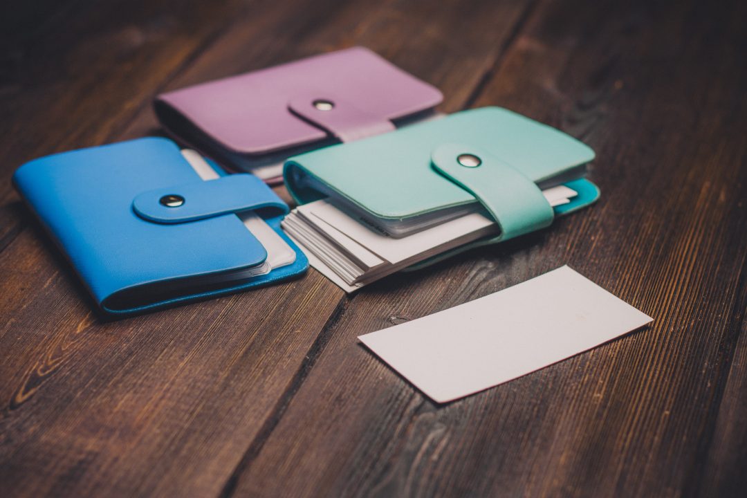 Leather business card cases in colors of light blue, purple, and teal. against a woodgrain background. A blank white business card is positioned in front of them