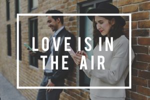 'Love is in the air' is written in white inside a square white border - in the background a nicely-dressed man and woman lean against a brick wall looking through their cell phones.