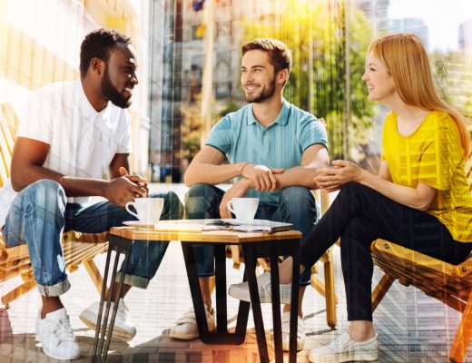 An African American male, a white male, and a strawberry blonde female, all in their early thirties, are sitting in wooden chairs and drinking coffee at an outdoor cafe while smiling and talking.