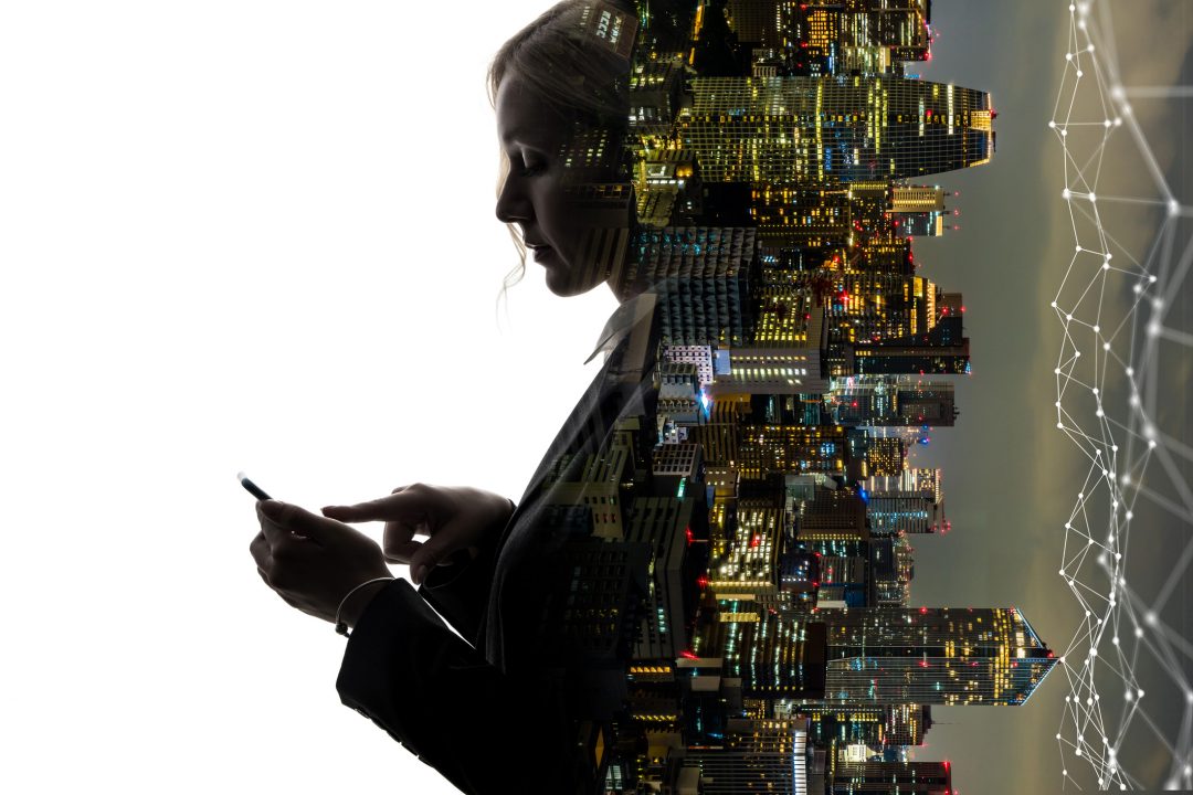 Attractive woman in business clothes types on her phone and leans against a city at night that has been inverted by 90 degrees.