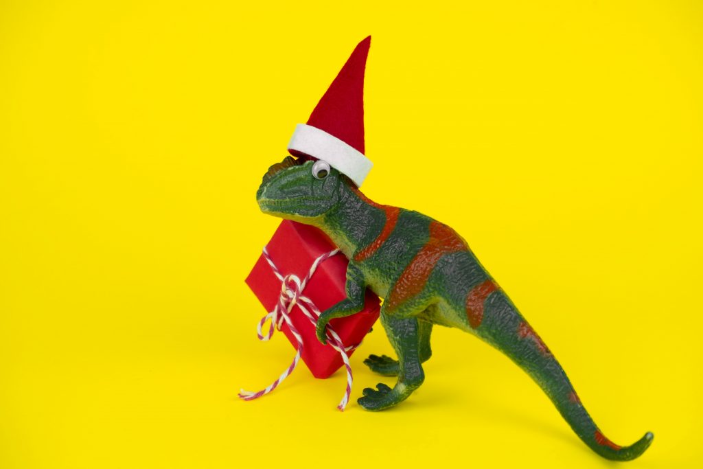funny green dinosaur toy in little santa claus hat with red gift box  on vibrant yellow background