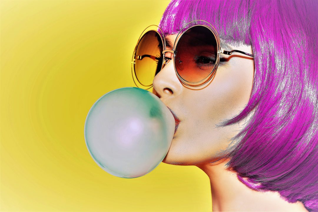 Pop art woman portrait wearing purple wig. Blow a blue bubble chewing gum. Olive background.