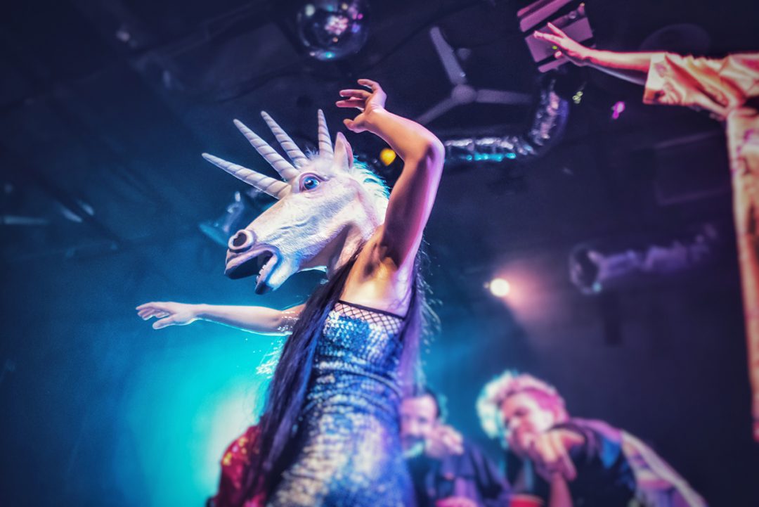 Human with a unicorn head dancing at a concert inside a nightclub.