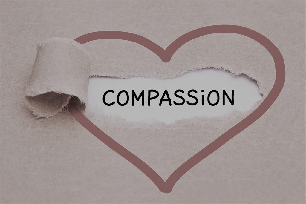 Handwritten word Compassion appearing behind ripped red heart on brown paper.