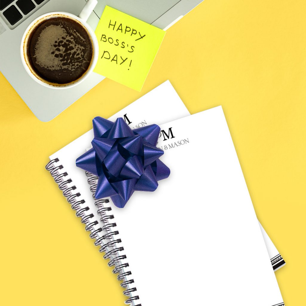 Post-it Note on yellow desk with coffee and laptop computer that says Happy Boss's Day next to a blank notebook with a blue ribbon on it.